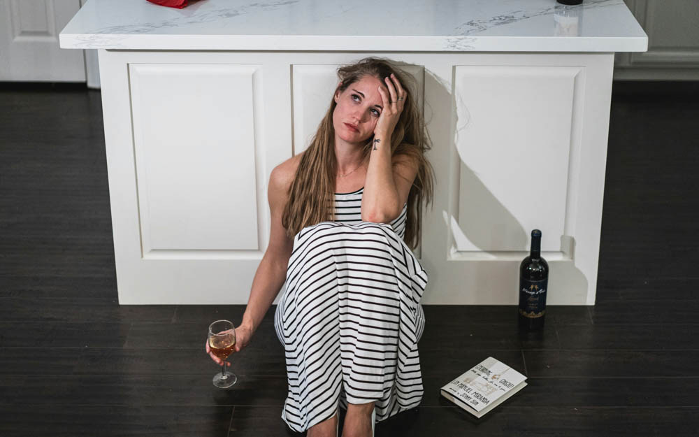 Photo of a drunk woman with messy hair sitting on the ground of her home. She has a bottle of wine next to her and a wine glass in hand.