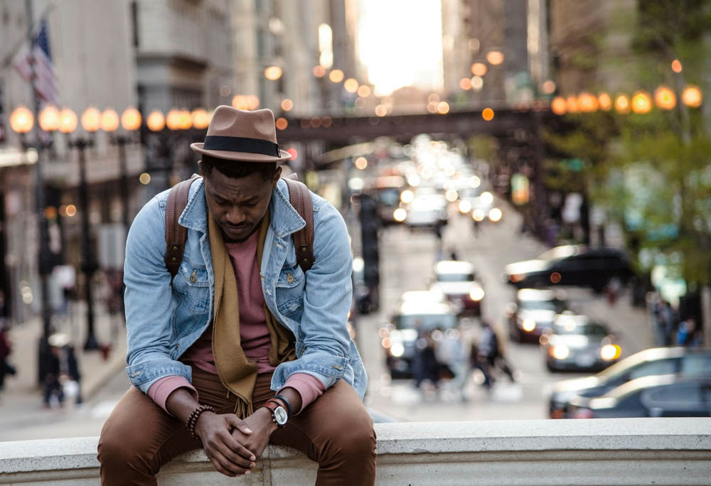 Photo of a man sitting on a balcony. A busy inner city street with people and cars in the background. He looks sad and lonely.