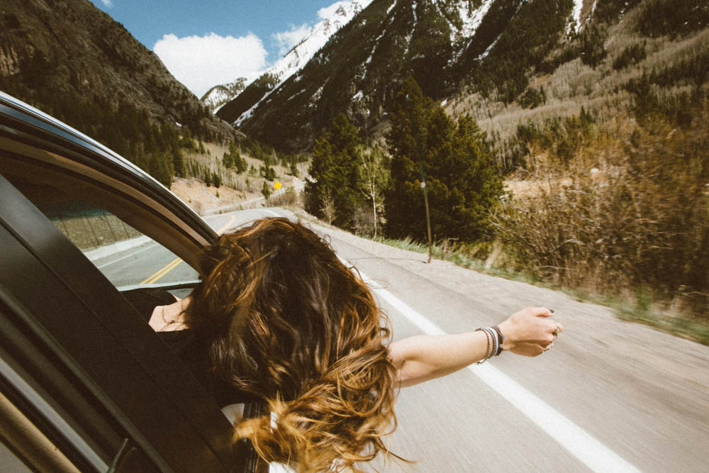 Photo of a woman with her head and arm hanging out of a car as it is driving down the road.