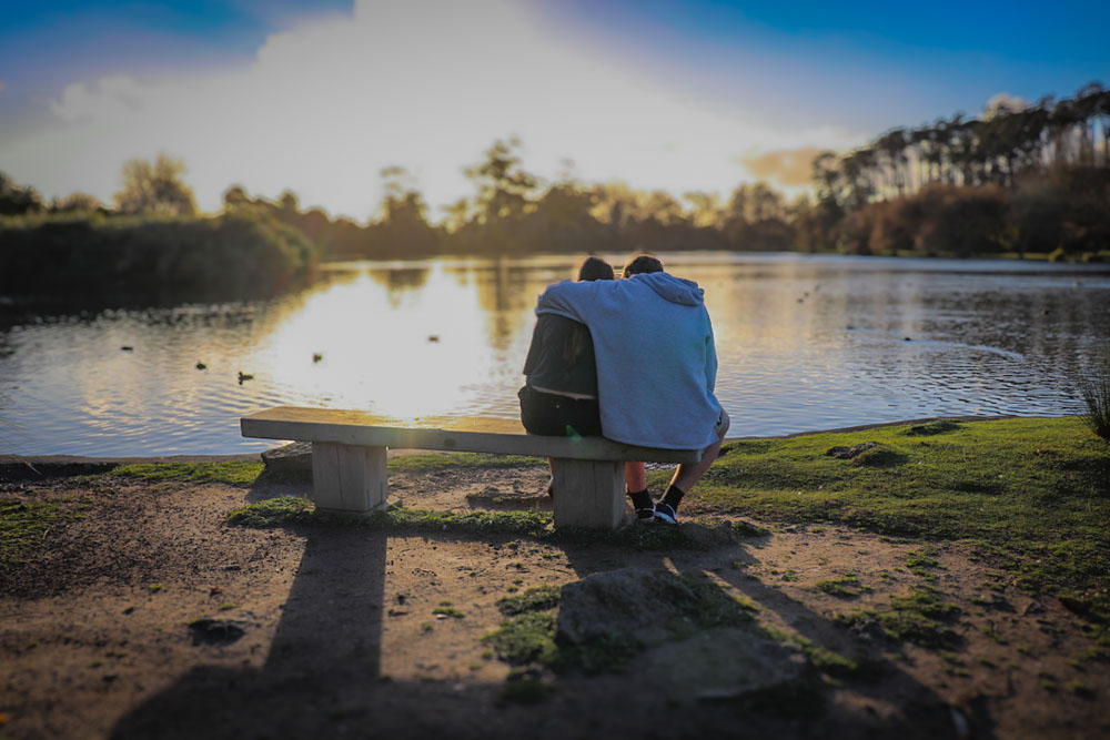 Photo at sunset of a couple sitting by a lake on a park bench cuddling.
