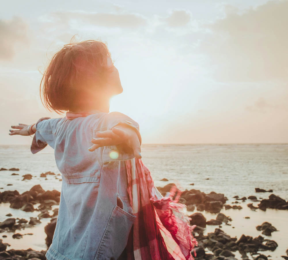 Photo of a woman standing on a beach on a sunny morning. She is looking out at the ocean with her arms out by her side, symbolising freedom.