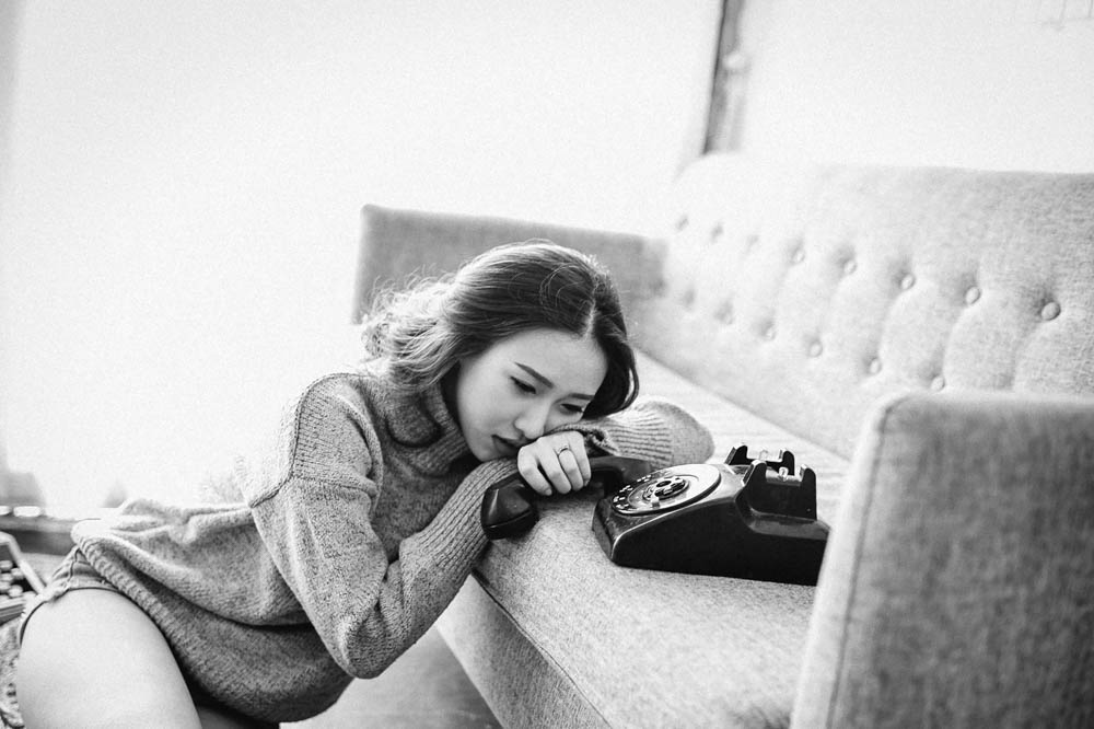 Photo of woman at home, sitting on the floor, her upper body leaning on a sofa, with a vintage phone handle in her hand. She looks distressed.