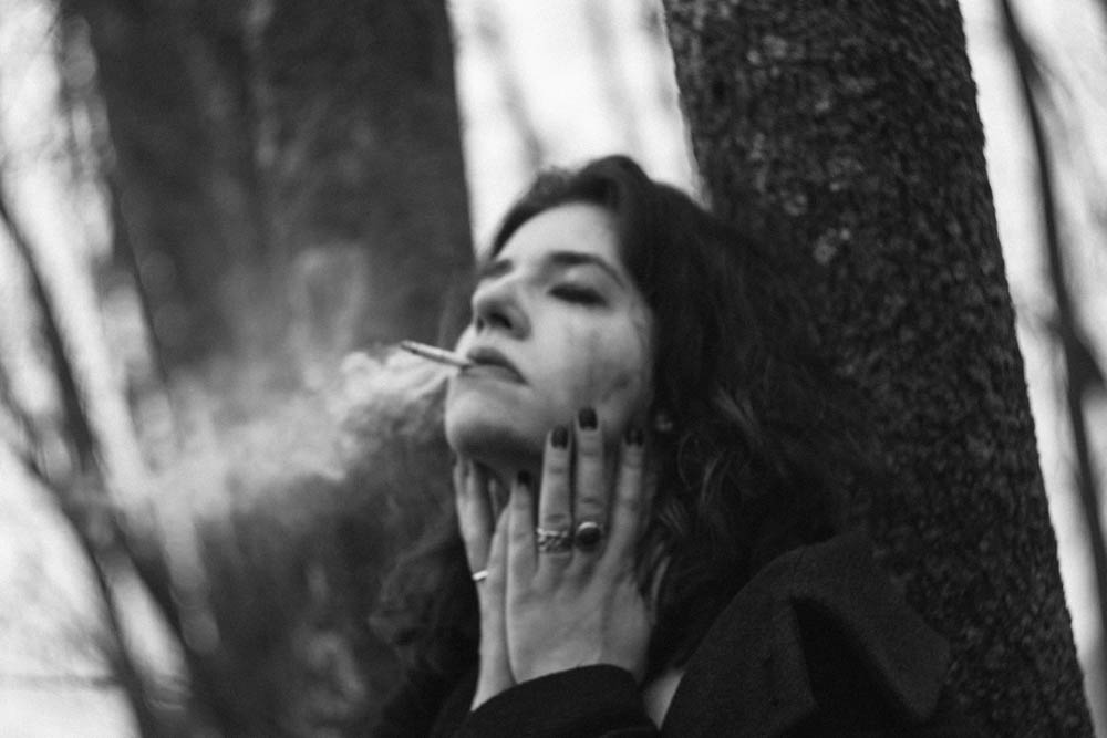 Photo of a young woman leaning against a tree, smoking and pulling at her face in frustration.
