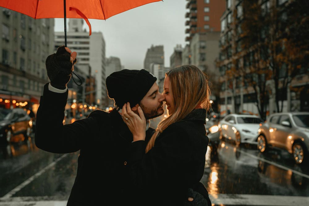 Photo of a man and woman kissing in the middle of a busy street, under an umbrella on a rainy day. Signs He's The One For You. 
