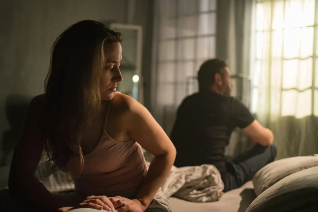Woman sitting on a bed looking distressed while her partner sits facing away towards a window, illustrating emotional distance in a troubled relationship.